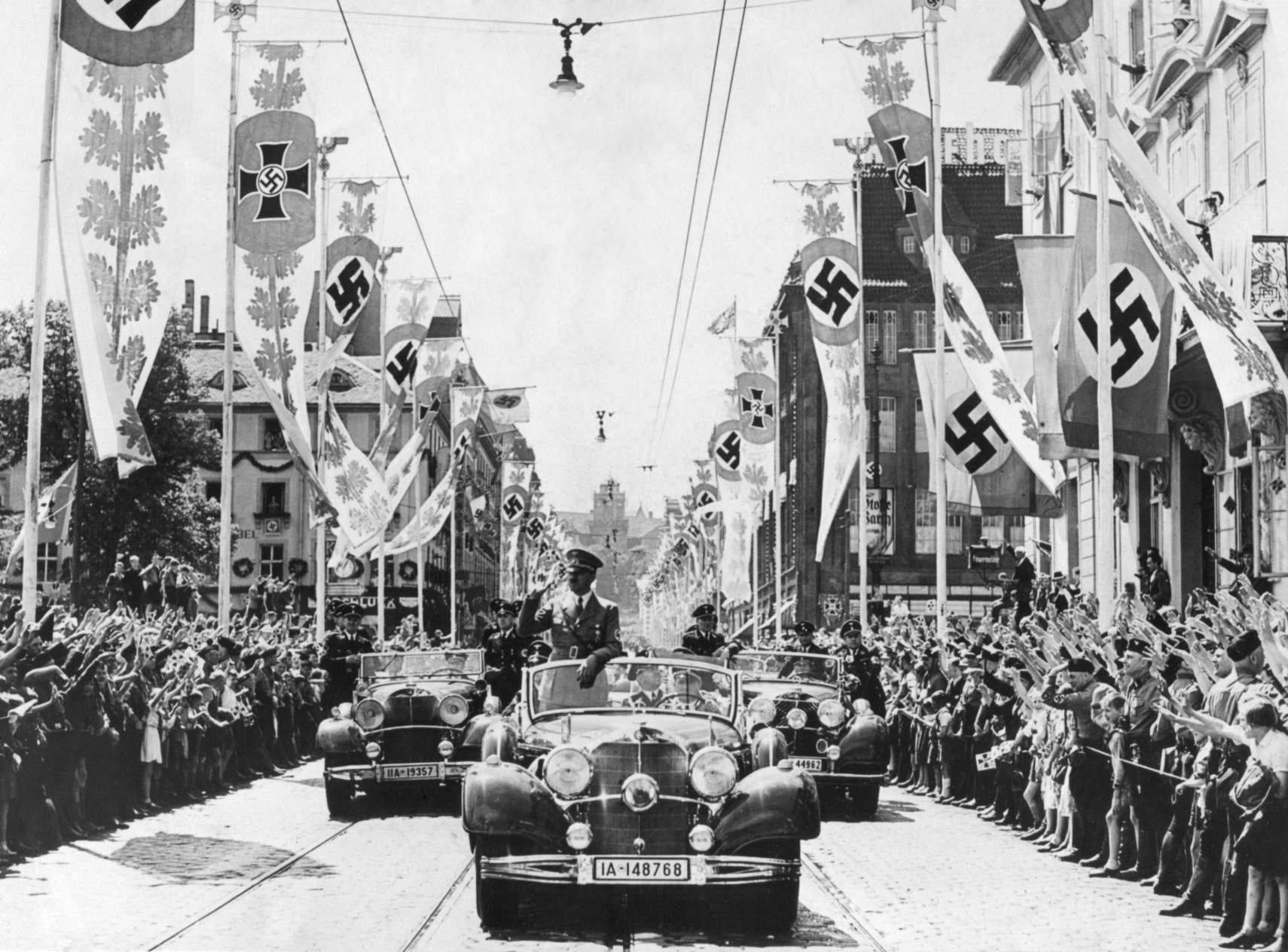 阿道夫·希特勒（Adolf Hitler）在遊行隊伍的前頭從他的車上向人群揮手致意。 街道上裝飾著各種萬字橫幅。 鈣。 1934-38 年。 希特勒選擇萬字符作為他們的標誌有一個方便但虛假的理由。 它在第二個千年被印度的雅利安游牧民族使用。 在納粹理論中，雅利安人有德國血統，希特勒得出結論，萬字符永遠是反猶太的。